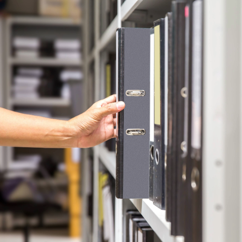 A person pulling a black lever arch file from a shelf filled with similar binders in an office archive