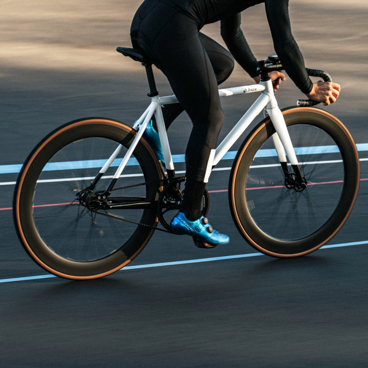 A cyclist riding a white road bike on a smooth track, wearing black cycling gear and blue shoes.